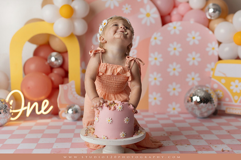 One Groovy Babe first birthday cake smash photography session at Studio 120 Photography in Ramona, California, featuring a baby girl in a pink and yellow retro daisy and disco themed set with balloons, a pink birthday cake, flower crown and vintage groovy van, followed by a rustic apple milk bath with red apples, florals and wooden accents.