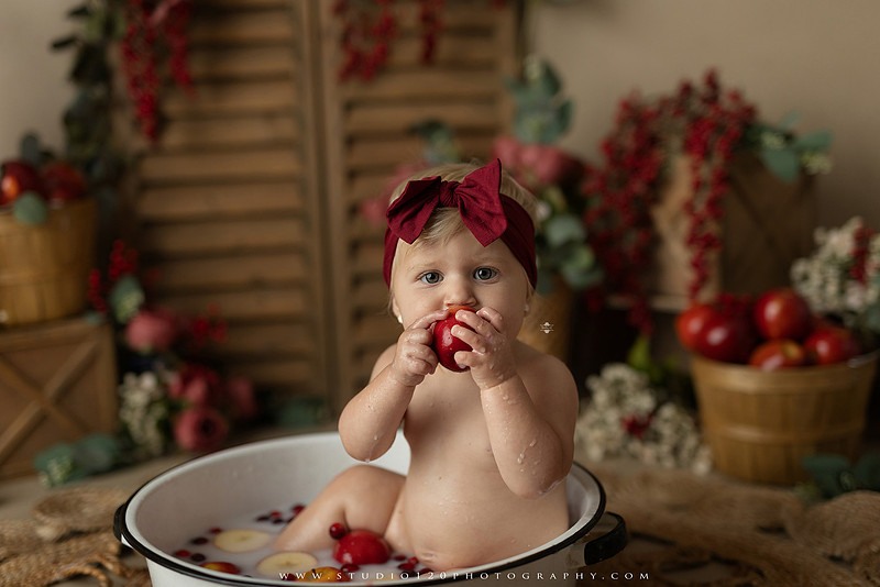 One Groovy Babe first birthday cake smash photography session at Studio 120 Photography in Ramona, California, featuring a baby girl in a pink and yellow retro daisy and disco themed set with balloons, a pink birthday cake, flower crown and vintage groovy van, followed by a rustic apple milk bath with red apples, florals and wooden accents.