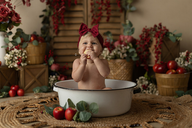 One Groovy Babe first birthday cake smash photography session at Studio 120 Photography in Ramona, California, featuring a baby girl in a pink and yellow retro daisy and disco themed set with balloons, a pink birthday cake, flower crown and vintage groovy van, followed by a rustic apple milk bath with red apples, florals and wooden accents.
