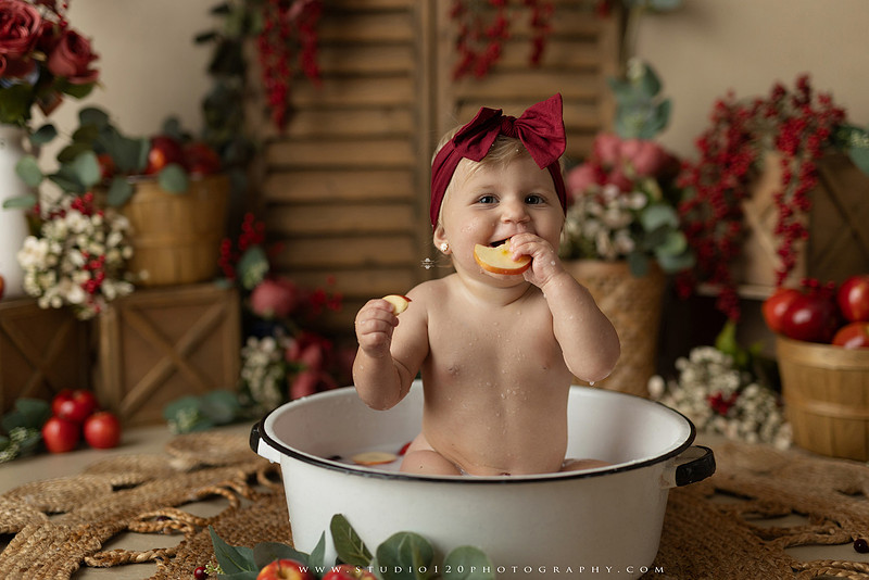 One Groovy Babe first birthday cake smash photography session at Studio 120 Photography in Ramona, California, featuring a baby girl in a pink and yellow retro daisy and disco themed set with balloons, a pink birthday cake, flower crown and vintage groovy van, followed by a rustic apple milk bath with red apples, florals and wooden accents.