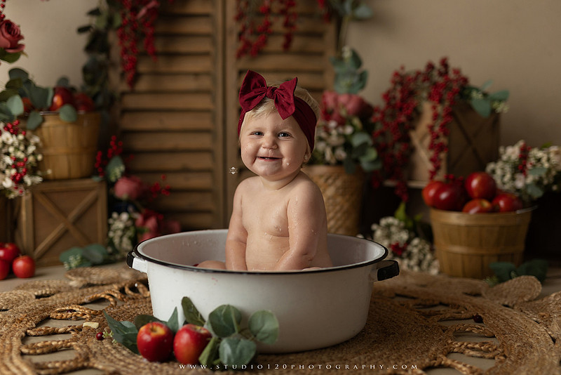 One Groovy Babe first birthday cake smash photography session at Studio 120 Photography in Ramona, California, featuring a baby girl in a pink and yellow retro daisy and disco themed set with balloons, a pink birthday cake, flower crown and vintage groovy van, followed by a rustic apple milk bath with red apples, florals and wooden accents.