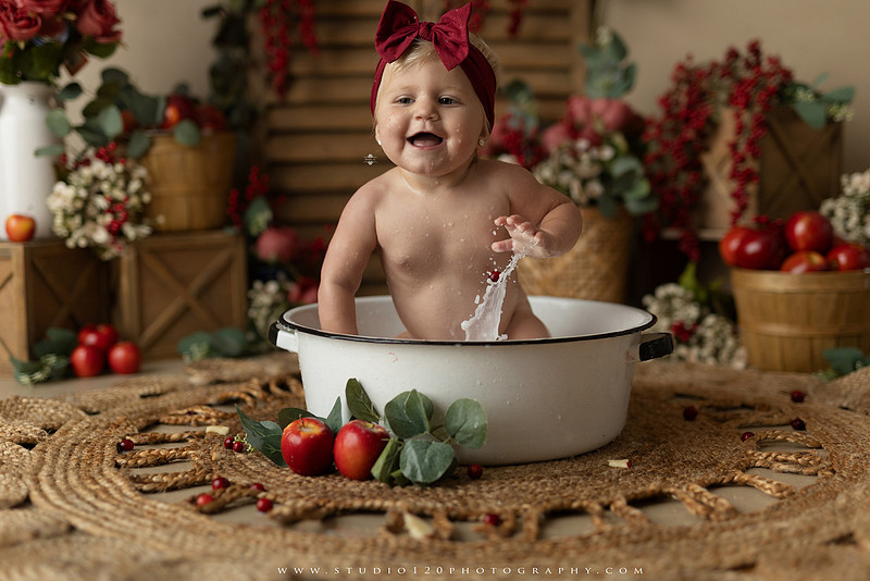 One Groovy Babe first birthday cake smash photography session at Studio 120 Photography in Ramona, California, featuring a baby girl in a pink and yellow retro daisy and disco themed set with balloons, a pink birthday cake, flower crown and vintage groovy van, followed by a rustic apple milk bath with red apples, florals and wooden accents.