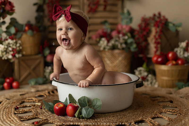 One Groovy Babe first birthday cake smash photography session at Studio 120 Photography in Ramona, California, featuring a baby girl in a pink and yellow retro daisy and disco themed set with balloons, a pink birthday cake, flower crown and vintage groovy van, followed by a rustic apple milk bath with red apples, florals and wooden accents.