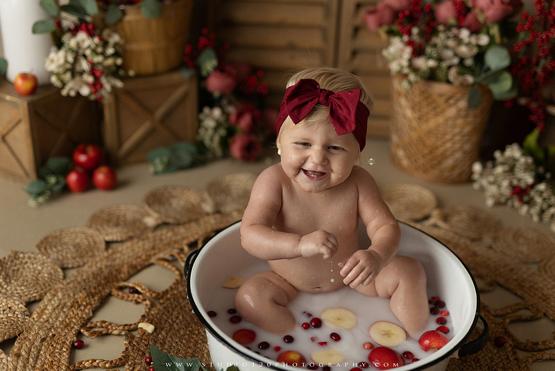 One Groovy Babe first birthday cake smash photography session at Studio 120 Photography in Ramona, California, featuring a baby girl in a pink and yellow retro daisy and disco themed set with balloons, a pink birthday cake, flower crown and vintage groovy van, followed by a rustic apple milk bath with red apples, florals and wooden accents.