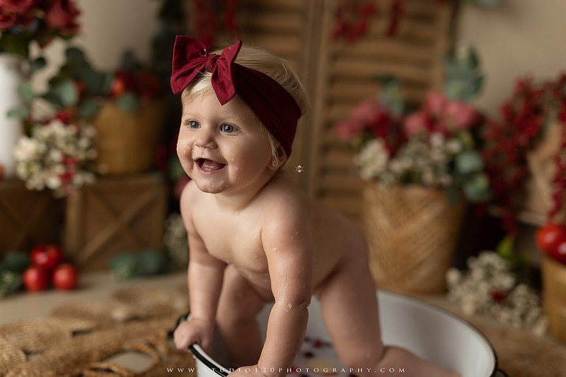 One Groovy Babe first birthday cake smash photography session at Studio 120 Photography in Ramona, California, featuring a baby girl in a pink and yellow retro daisy and disco themed set with balloons, a pink birthday cake, flower crown and vintage groovy van, followed by a rustic apple milk bath with red apples, florals and wooden accents.