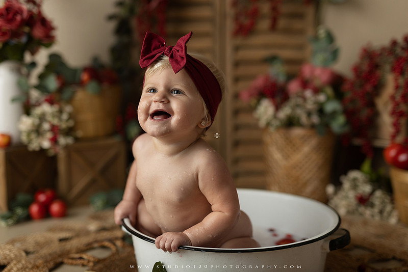 One Groovy Babe first birthday cake smash photography session at Studio 120 Photography in Ramona, California, featuring a baby girl in a pink and yellow retro daisy and disco themed set with balloons, a pink birthday cake, flower crown and vintage groovy van, followed by a rustic apple milk bath with red apples, florals and wooden accents.