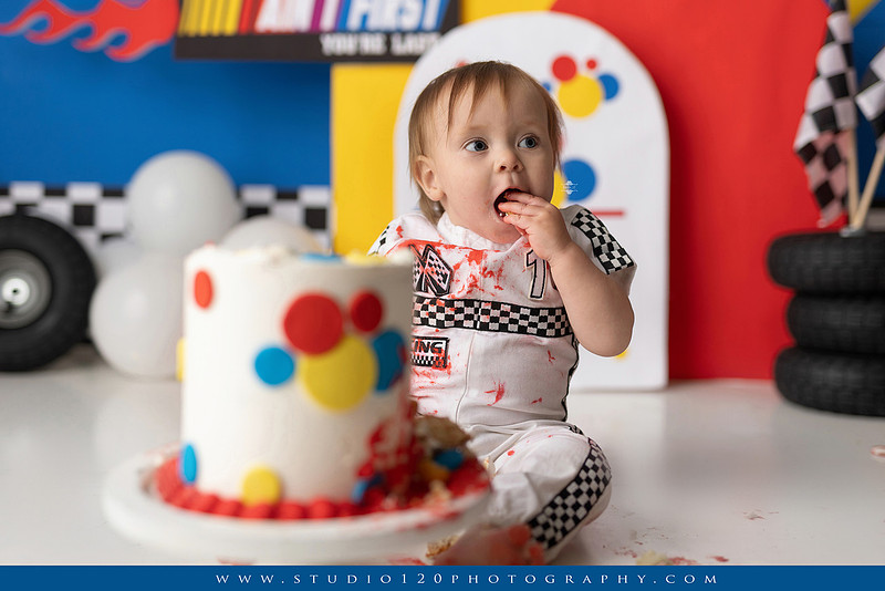 Baby boy celebrating his first birthday with a Talladega Nights-inspired racing cake smash, featuring a colorful race car theme, checkered flags, racing tires, bright red, yellow and blue décor, and a custom birthday cake at Studio 120 Photography in Ramona, California.