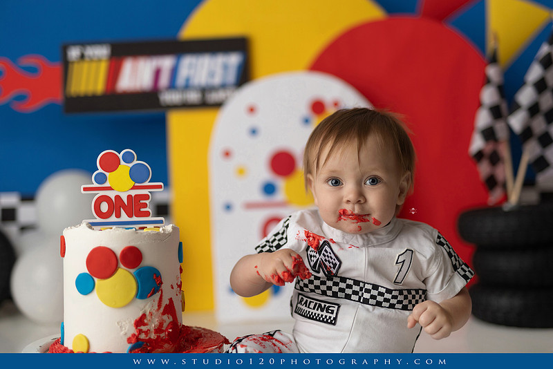 Baby boy celebrating his first birthday with a Talladega Nights-inspired racing cake smash, featuring a colorful race car theme, checkered flags, racing tires, bright red, yellow and blue décor, and a custom birthday cake at Studio 120 Photography in Ramona, California.