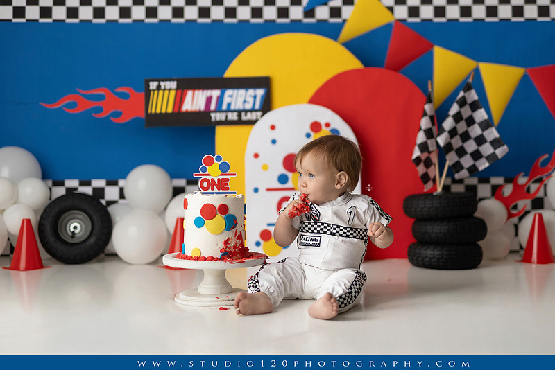 Baby boy celebrating his first birthday with a Talladega Nights-inspired racing cake smash, featuring a colorful race car theme, checkered flags, racing tires, bright red, yellow and blue décor, and a custom birthday cake at Studio 120 Photography in Ramona, California.