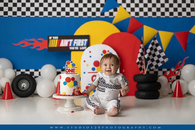 Baby boy celebrating his first birthday with a Talladega Nights-inspired racing cake smash, featuring a colorful race car theme, checkered flags, racing tires, bright red, yellow and blue décor, and a custom birthday cake at Studio 120 Photography in Ramona, California.