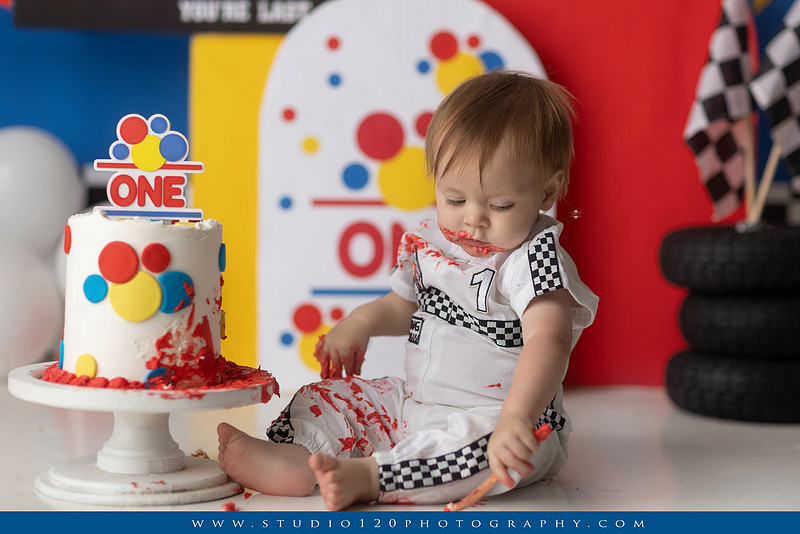 Baby boy celebrating his first birthday with a Talladega Nights-inspired racing cake smash, featuring a colorful race car theme, checkered flags, racing tires, bright red, yellow and blue décor, and a custom birthday cake at Studio 120 Photography in Ramona, California.