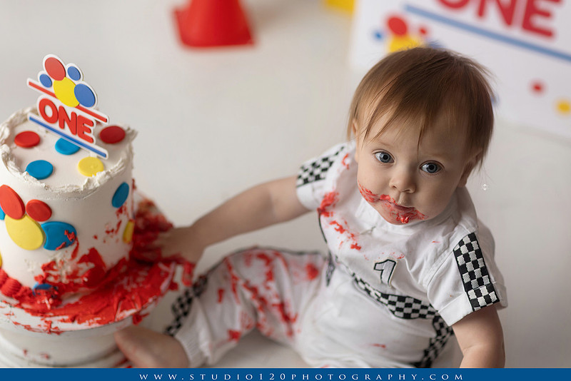 Baby boy celebrating his first birthday with a Talladega Nights-inspired racing cake smash, featuring a colorful race car theme, checkered flags, racing tires, bright red, yellow and blue décor, and a custom birthday cake at Studio 120 Photography in Ramona, California.