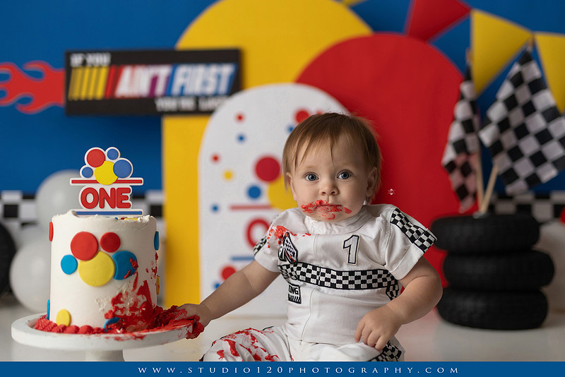 Baby boy celebrating his first birthday with a Talladega Nights-inspired racing cake smash, featuring a colorful race car theme, checkered flags, racing tires, bright red, yellow and blue décor, and a custom birthday cake at Studio 120 Photography in Ramona, California.
