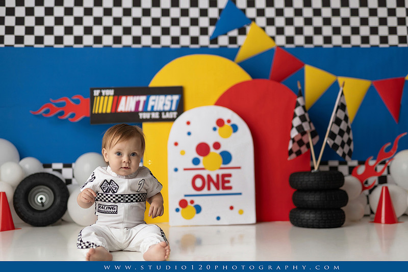 Baby boy celebrating his first birthday with a Talladega Nights-inspired racing cake smash, featuring a colorful race car theme, checkered flags, racing tires, bright red, yellow and blue décor, and a custom birthday cake at Studio 120 Photography in Ramona, California.