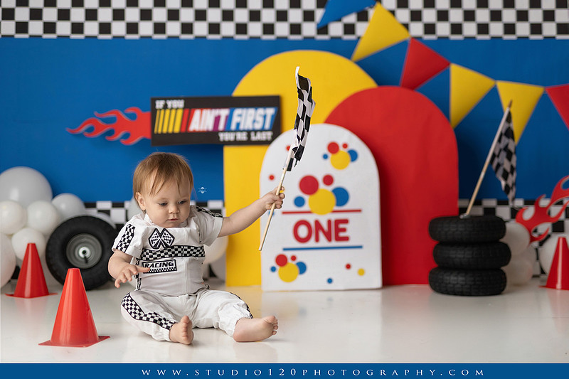Baby boy celebrating his first birthday with a Talladega Nights-inspired racing cake smash, featuring a colorful race car theme, checkered flags, racing tires, bright red, yellow and blue décor, and a custom birthday cake at Studio 120 Photography in Ramona, California.