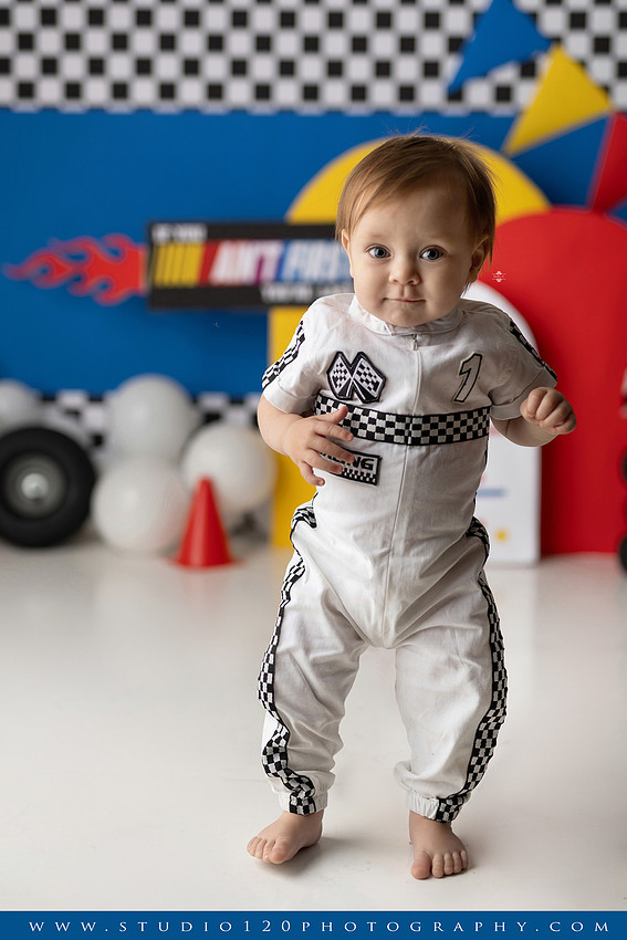 Baby boy celebrating his first birthday with a Talladega Nights-inspired racing cake smash, featuring a colorful race car theme, checkered flags, racing tires, bright red, yellow and blue décor, and a custom birthday cake at Studio 120 Photography in Ramona, California.
