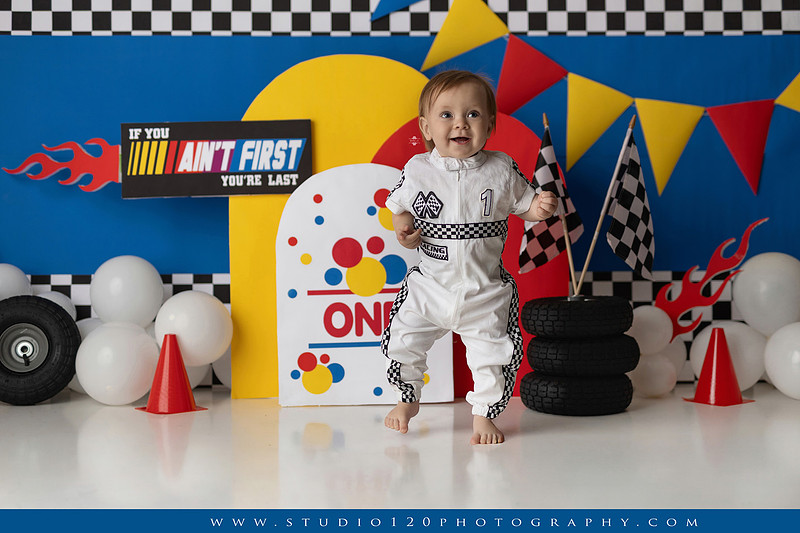 Baby boy celebrating his first birthday with a Talladega Nights-inspired racing cake smash, featuring a colorful race car theme, checkered flags, racing tires, bright red, yellow and blue décor, and a custom birthday cake at Studio 120 Photography in Ramona, California.