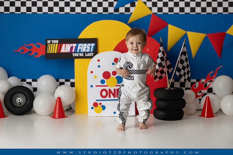 Baby boy celebrating his first birthday with a Talladega Nights-inspired racing cake smash, featuring a colorful race car theme, checkered flags, racing tires, bright red, yellow and blue décor, and a custom birthday cake at Studio 120 Photography in Ramona, California.