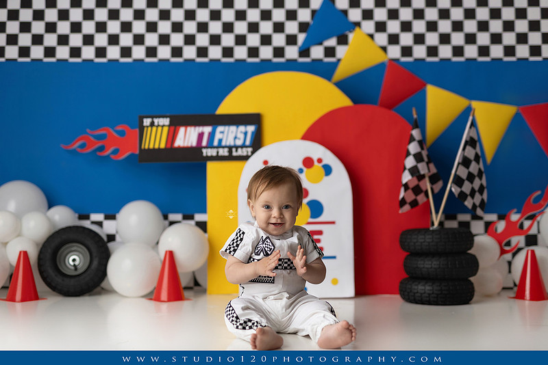 Baby boy celebrating his first birthday with a Talladega Nights-inspired racing cake smash, featuring a colorful race car theme, checkered flags, racing tires, bright red, yellow and blue décor, and a custom birthday cake at Studio 120 Photography in Ramona, California.