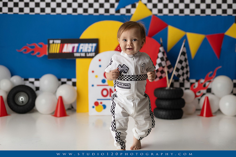 Baby boy celebrating his first birthday with a Talladega Nights-inspired racing cake smash, featuring a colorful race car theme, checkered flags, racing tires, bright red, yellow and blue décor, and a custom birthday cake at Studio 120 Photography in Ramona, California.