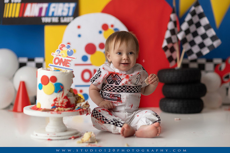 Baby boy celebrating his first birthday with a Talladega Nights-inspired racing cake smash, featuring a colorful race car theme, checkered flags, racing tires, bright red, yellow and blue décor, and a custom birthday cake at Studio 120 Photography in Ramona, California.