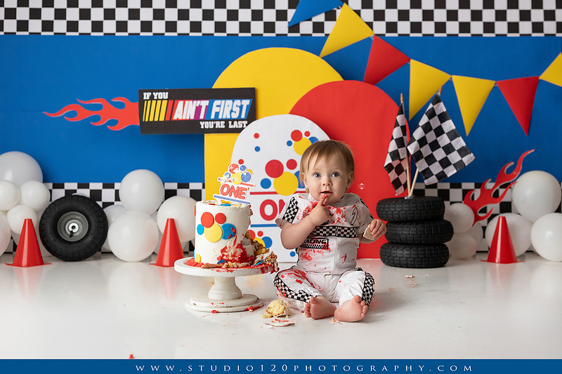 Baby boy celebrating his first birthday with a Talladega Nights-inspired racing cake smash, featuring a colorful race car theme, checkered flags, racing tires, bright red, yellow and blue décor, and a custom birthday cake at Studio 120 Photography in Ramona, California.