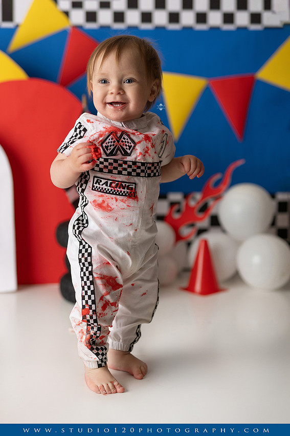Baby boy celebrating his first birthday with a Talladega Nights-inspired racing cake smash, featuring a colorful race car theme, checkered flags, racing tires, bright red, yellow and blue décor, and a custom birthday cake at Studio 120 Photography in Ramona, California.