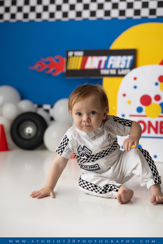 Baby boy celebrating his first birthday with a Talladega Nights-inspired racing cake smash, featuring a colorful race car theme, checkered flags, racing tires, bright red, yellow and blue décor, and a custom birthday cake at Studio 120 Photography in Ramona, California.