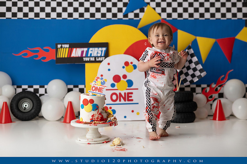 Baby boy celebrating his first birthday with a Talladega Nights-inspired racing cake smash, featuring a colorful race car theme, checkered flags, racing tires, bright red, yellow and blue décor, and a custom birthday cake at Studio 120 Photography in Ramona, California.