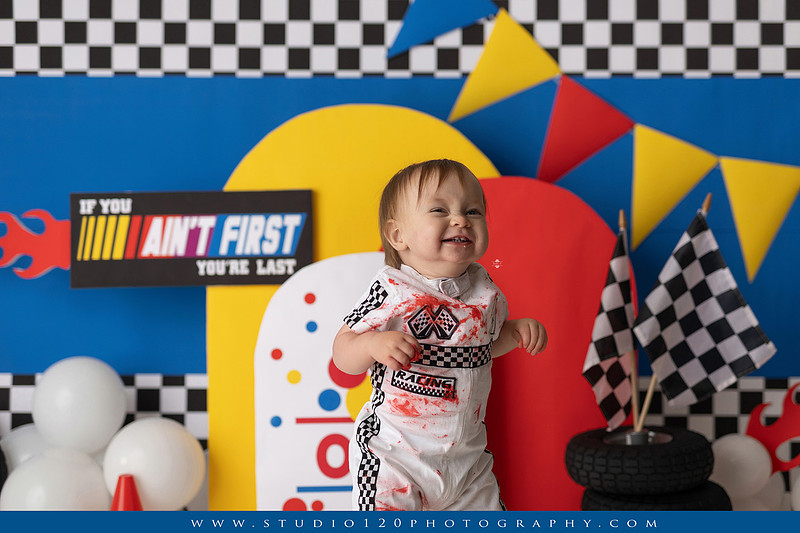 Baby boy celebrating his first birthday with a Talladega Nights-inspired racing cake smash, featuring a colorful race car theme, checkered flags, racing tires, bright red, yellow and blue décor, and a custom birthday cake at Studio 120 Photography in Ramona, California.